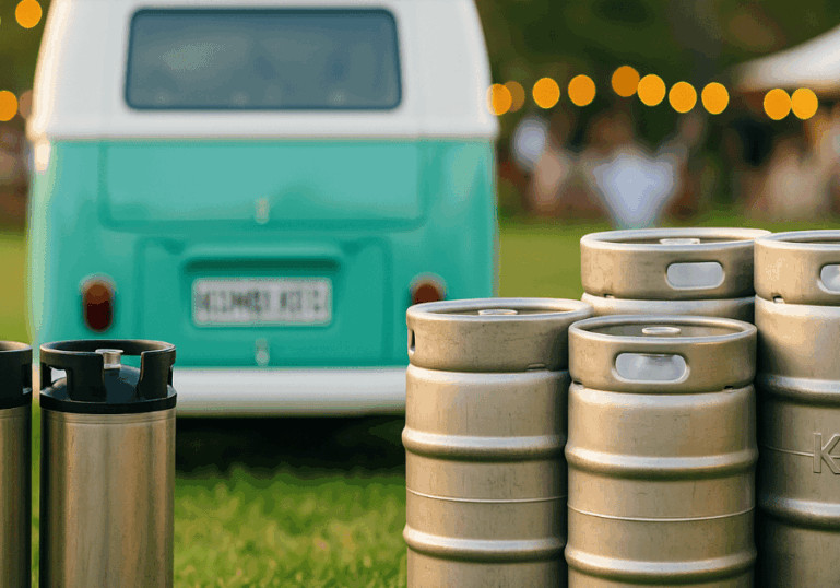 Stainless steel kegs arranged on grass with a teal and white Kombi Keg van blurred in the background at a festive outdoor event.